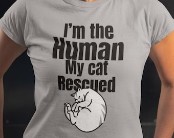 May include: A light gray t-shirt with the text "I'm the Human My cat Rescued" in black. Below the text is a white illustration of a curled-up cat. The shirt is short-sleeved.