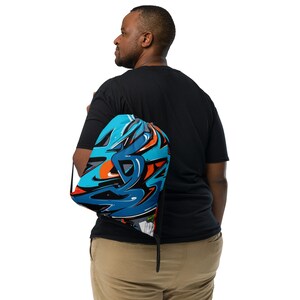 Puede incluir: Una mochila con cordón azul con un vibrante diseño de graffiti en tonos de azul, naranja y negro. La bolsa se lleva sobre el hombro, mostrando la colorida obra de arte abstracta. El diseño de la bolsa es llamativo y moderno.