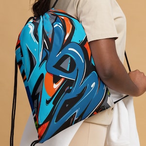 Puede incluir: Una mochila con cordón azul con un diseño de estilo grafiti. El diseño presenta formas azules, naranjas y negras en un fondo azul claro. La bolsa tiene cordones negros y se lleva sobre el hombro.