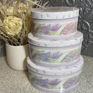 Könnte beinhalten: Drei runde, stapelbare, metallene Aufbewahrungsdosen mit einem Lavendel-Blumenmuster. Die Dosen sind weiß mit einem lila und grünen Blumenmuster und einem gelben Band.
