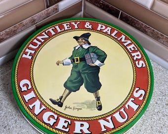 May include: A round green, red, and white tin with the words "Huntley & Palmers Ginger Nuts" and a cartoon illustration of a man in a green suit holding a box of ginger nuts. The text "John Ginger" is written below the illustration.