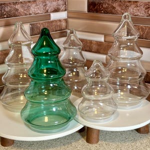 Pode incluir: Conjunto de cinco jarros de doces de vidro em forma de árvore de Natal. Quatro são de vidro transparente e um é de vidro verde.