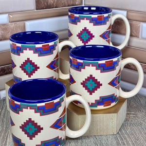 May include: Four ceramic mugs with a cream-colored background and a Southwestern geometric pattern in teal, red, and blue. The mugs have a blue interior and a white handle. The mugs are displayed on a wooden surface.
