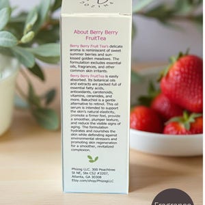 May include: A white box of Berry Berry FruitTea serum with text describing the product's benefits. The box is next to a bowl of fresh strawberries and a sprig of greenery. The label says "Fragrance-free".