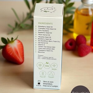May include: A white product box with the word "INGREDIENTS" and a list of ingredients, including Strawberry Seed Oil and Bakuchiol. The box is next to a cut strawberry and a bottle of oil. The text "Bakuchiol Inside" is on a dark circle.