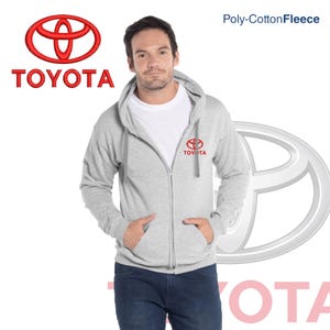 Puede incluir: Sudadera con capucha gris claro con cremallera y el logotipo de Toyota rojo en el pecho y un logotipo más grande a la izquierda. La palabra "TOYOTA" está bordada en rojo. El texto "Poly-Cotton Fleece" está en la esquina superior derecha.