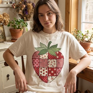 May include: A cream-coloured short-sleeve t-shirt featuring a large, central graphic of a strawberry constructed from a patchwork of red and white fabrics. The strawberry design includes various patterns like gingham, hearts, and small floral motifs.