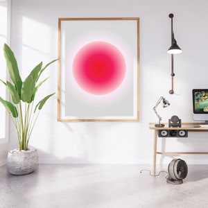 Può includere: Una stampa incorniciata di un cerchio sfumato rosa e bianco su sfondo bianco. La stampa è appesa a una parete bianca sopra una scrivania con una lampada, un ventilatore e un altoparlante.