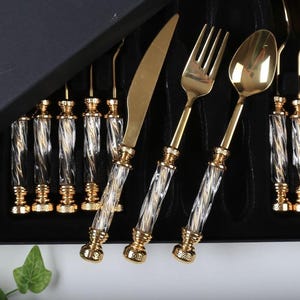 Könnte beinhalten: Ein goldfarbenes Besteckset mit transparenten und weiß gemusterten Griffen, präsentiert in einer schwarzen Box. Das Set enthält Messer, Gabeln und Löffel. Die Griffe haben ein dekoratives, gedrehtes Design. Die Box ist geöffnet.