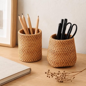 Può includere: Due portamatite cilindrici in rattan intrecciato di colore beige chiaro. Un contenitore ospita quattro matite in legno, mentre l'altro contiene penne nere e un paio di forbici nere. I contenitori presentano un motivo a intreccio fitto.