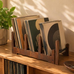 Op de afbeelding: Een donkerbruine houten vinylplatenhouder met meerdere platen met diverse grafische hoezen. Het rek is voorzien van verticale verdelers en een horizontale zwarte metalen steunstang om de platen rechtop en geordend te houden.