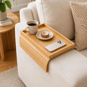 May include: A rectangular bamboo sofa arm tray with a flexible, slatted design that drapes over the armrest. The flat top surface holds a ceramic mug filled with dark coffee, a small plate with a pastry, and a silver smartphone.