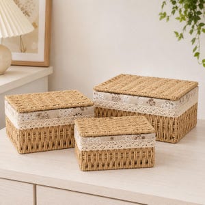 Pode incluir: Três cestos de armazenamento retangulares com tampas, feitos de material trançado em tom bronzeado. Cada cesto apresenta uma faixa de tecido bege decorada com um padrão floral branco e um delicado acabamento em renda branca no topo.