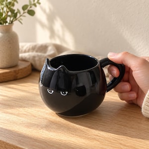 Puede incluir: Una taza de cerámica negra brillante con forma de cabeza de gato, orejas puntiagudas y ojos impresos en blanco. La taza presenta un cuerpo redondeado y un asa curva en el lado derecho. La superficie es lisa y reflectante con brillos de luz.