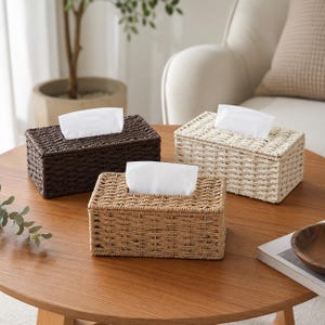 May include: Three rectangular tissue box covers made of woven material, each featuring a top opening with a white tissue pulled through. The set includes one dark brown, one light beige, and one natural tan woven box cover for standard tissues.