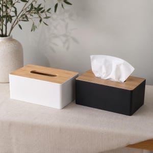 以下が含まれることがあります： 白いベースと黒いベースの2つの長方形のティッシュボックスカバー。どちらも中央に楕円形の開口部がある明るい色の木製蓋が付いています。黒いボックスには、上部の開口部から白いティッシュが出ています。
