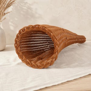 Könnte beinhalten: Ein brauner, füllhornförmiger Korb aus geflochtenem Material. Der Korb besitzt eine weite, offene kreisförmige Öffnung mit einem geflochtenen Rand, der sich zu einem schmalen, länglichen Ende hin verjüngt und ein gleichmäßiges Flechtmuster aufweist.