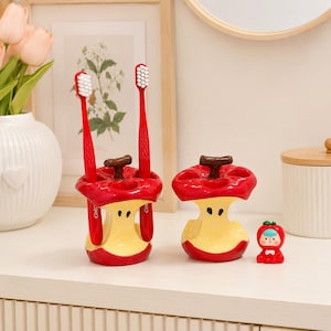 May include: Two ceramic toothbrush holders shaped like apple cores with red skin and pale yellow flesh, featuring brown stems. One holder contains two red toothbrushes. A small red apple-shaped figurine with a face sits next to the holders.