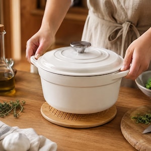 Peut inclure: Une cocotte en fonte émaillée blanche avec un couvercle assorti et un bouton en métal argenté. La marmite ronde possède deux poignées latérales et repose sur un dessous de plat en bois circulaire à surface rainurée, au design épuré.