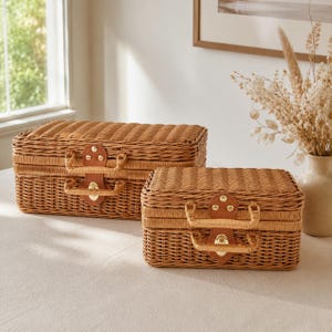 May include: Two rectangular woven wicker suitcases in a natural light brown finish. Each case features a hinged lid, a woven handle, and a central tan leather-like latch plate secured with gold-toned metal rivets and a polished gold-toned clasp.