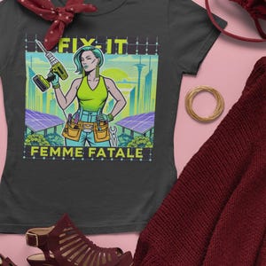 May include: A black t-shirt with a graphic of a woman holding a drill, the words "FIX IT" and "FEMME FATALE". The woman is wearing a green top and tool belt. The background features a cityscape and solar panels.