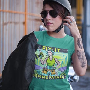 May include: A teal t-shirt with the words "FIX IT" and "FEMME FATALE" printed on it. The graphic features a woman holding a drill, with a cityscape and solar panels in the background. The person in the image is wearing a black helmet and sunglasses.