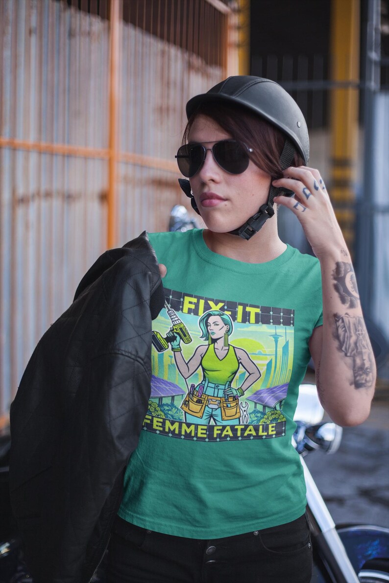 May include: A teal t-shirt with the words "FIX IT" and "FEMME FATALE" printed on it. The graphic features a woman holding a drill, with a cityscape and solar panels in the background. The person in the image is wearing a black helmet and sunglasses.