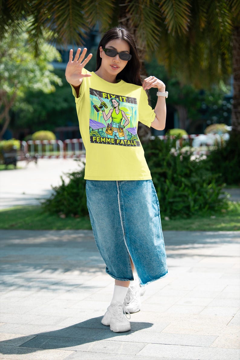 May include: Yellow t-shirt with the text "FIX IT" and "FEMME FATALE" featuring a cartoon woman holding a drill. The woman is wearing a green top and tool belt. The model is wearing a denim skirt and white trainers.