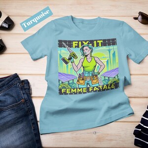 May include: A turquoise t-shirt featuring a graphic of a woman with a drill, the text "FIX IT" and "FEMME FATALE". The image also includes a pair of blue jeans, black and white shoes, sunglasses, and a striped shirt.