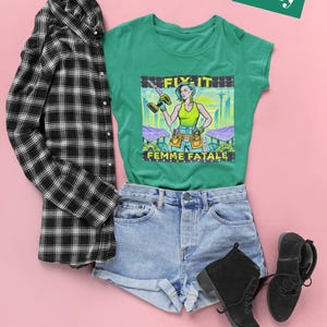 May include: A green t-shirt with a graphic of a woman holding a drill, the text "FIX IT FEMME FATALE". The shirt is styled with denim shorts, black boots, and a black and white checked shirt. A green rectangle with the name "Kelly" is in the top right corner.
