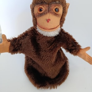 May include: A vintage brown plush monkey puppet with a tan face, large brown eyes, and a small red nose. The puppet has a white fur collar and outstretched arms. The monkey's fur is a deep brown colour.