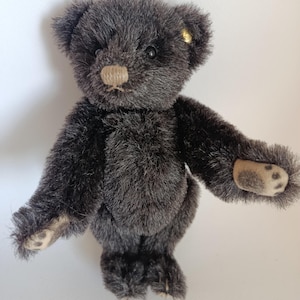 Puede incluir: Un pequeño oso de peluche gris oscuro con un hocico y almohadillas de las patas de color marrón claro. El oso tiene un diseño clásico con una apariencia ligeramente envejecida, y una pequeña etiqueta dorada es visible en su oreja. El oso está sobre una superficie blanca.