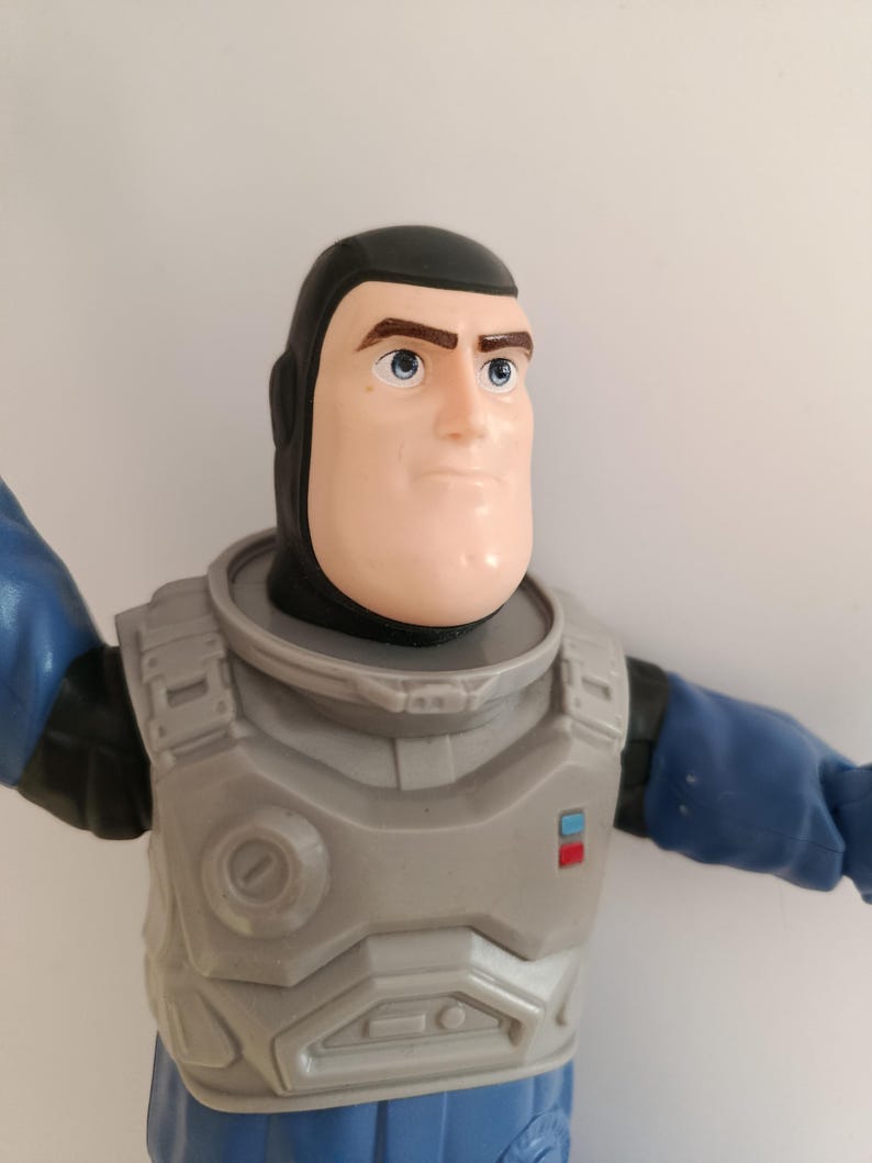 May include: A Buzz Lightyear action figure, featuring a light skin tone, black hair, and blue eyes. The figure wears a gray and blue space suit with a chest plate and blue sleeves. The toy is a collectible item.