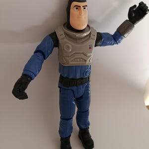 May include: A Buzz Lightyear action figure in a blue and gray space suit. The figure has a white face, black gloves, and black boots. The figure is standing with one arm raised.