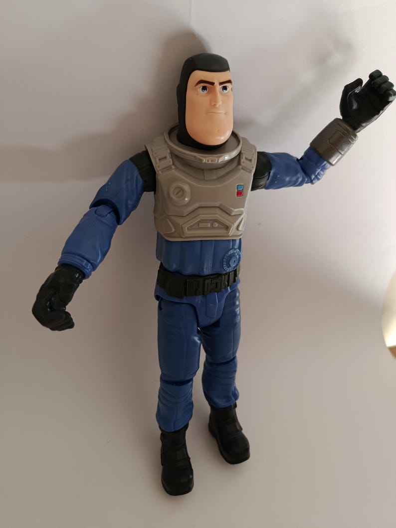 May include: A Buzz Lightyear action figure in a blue and gray space suit. The figure has a white face, black gloves, and black boots. The figure is standing with one arm raised.