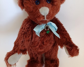 Puede incluir: Un oso de peluche marrón hecho a mano con ojos azules, hocico blanco y almohadillas grises. Tiene una cinta azul claro atada alrededor del cuello con un amuleto de herradura. El oso está de pie.