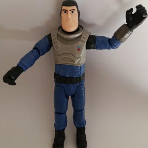 May include: A Buzz Lightyear action figure in a blue and gray space suit. The figure has a tan face, black hair, and black gloves. The figure's arms are outstretched, and it is standing on a white surface.