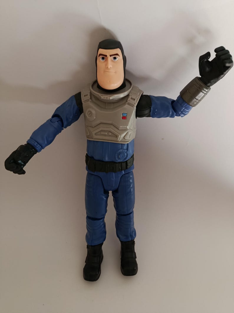 May include: A Buzz Lightyear action figure in a blue and gray space suit. The figure has a tan face, black hair, and black gloves. The figure's arms are outstretched, and it is standing on a white surface.