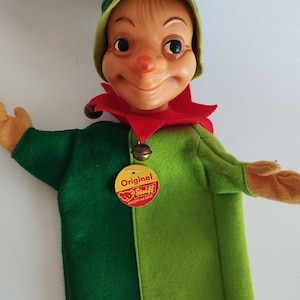 Puede incluir: Marionetas de mano de fieltro verde y amarillo con cara marrón y cuello rojo. La marioneta tiene un sombrero verde y una etiqueta que dice "Original Steiff Knopf im Ohr".