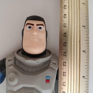 May include: A Buzz Lightyear action figure, featuring a gray and blue space suit and a stern expression. The figure is approximately 10 inches tall, with a ruler placed next to it for scale. The figure has a black helmet and blue eyes.