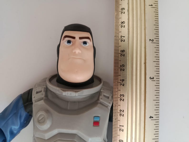 May include: A Buzz Lightyear action figure, featuring a gray and blue space suit and a stern expression. The figure is approximately 10 inches tall, with a ruler placed next to it for scale. The figure has a black helmet and blue eyes.