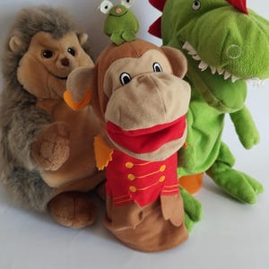 Puede incluir: Tres juguetes de peluche: un erizo, un mono títere con una chaqueta roja y un dinosaurio verde. El erizo es marrón y gris. El mono tiene una rana en la cabeza. El dinosaurio tiene púas rojas.