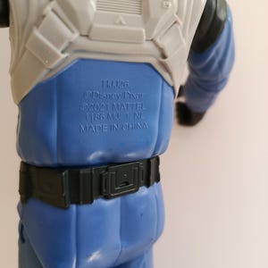 May include: A blue and white action figure with the text "HJJ26 @Disney/Pixar &copy;2021 MATTEL 1186 MJ. 1. NL. MADE IN CHINA". The figure has a black belt and blue pants.
