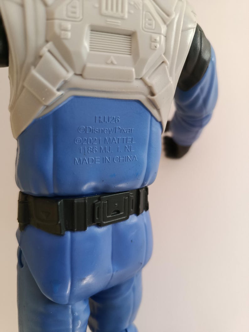 May include: A blue and white action figure with the text "HJJ26 @Disney/Pixar &copy;2021 MATTEL 1186 MJ. 1. NL. MADE IN CHINA". The figure has a black belt and blue pants.