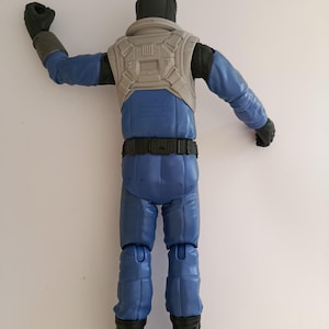 May include: A blue action figure of a character in a futuristic outfit. The figure has a black helmet, gloves, and boots. It wears a gray vest and a black belt. The figure's arms are outstretched, and it is posed against a white background.