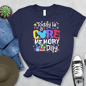 May include: Navy blue t-shirt with the text "Today is a Core Memory Day" in colorful letters and a Mickey Mouse head. The shirt also features cartoon characters. A pair of black and white sneakers and blue jeans are next to the shirt.