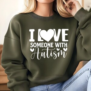 May include: Olive green sweatshirt with the text "I LOVE SOMEONE WITH Autism" in white. The word "LOVE" features a heart symbol. The sweatshirt has a crew neck and long sleeves.