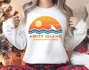 May include: White sweatshirt featuring a retro graphic of a sunset over ocean waves with a shark fin. The text "AMITY ISLAND 1975" is printed below the graphic in orange. A casual, comfortable top.
