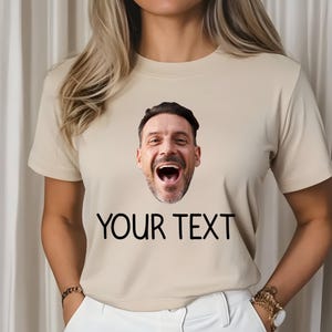 May include: Beige t-shirt with a printed image of a man's face and the text "YOUR TEXT" in black. The man's face is a close-up, showing him laughing with his mouth wide open. The shirt has short sleeves.