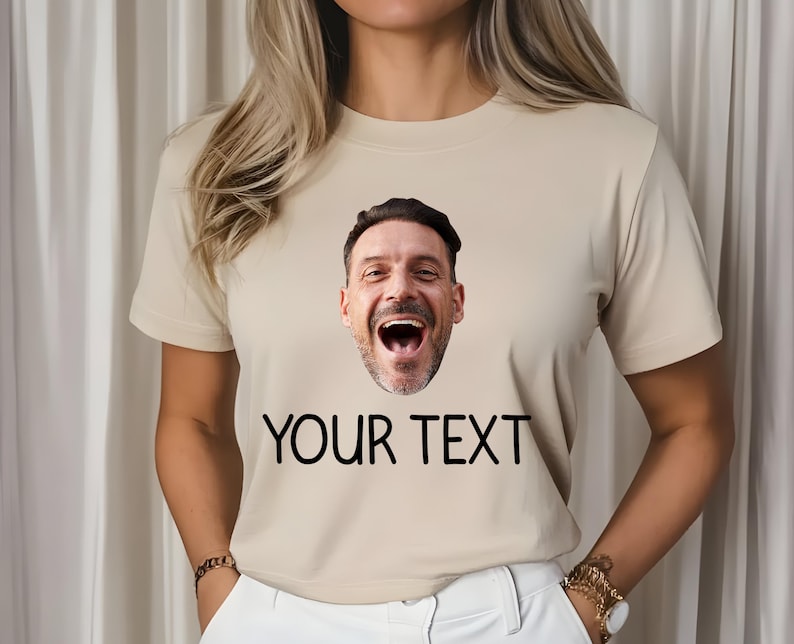 May include: Beige t-shirt with a printed image of a man's face and the text "YOUR TEXT" in black. The man's face is a close-up, showing him laughing with his mouth wide open. The shirt has short sleeves.